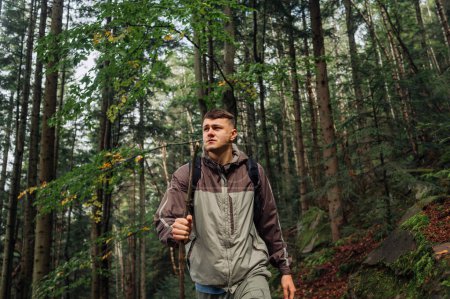 Photo for A handsome male hiker with a backpack and a stick in his hand is walking in the mountains through a dense forest with a serious face and looking away. Active tourism. - Royalty Free Image