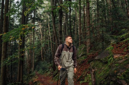 Photo for A handsome young man in casual clothes walks through the forest in the mountains with a serious face and looks away. Hiking trips alone. - Royalty Free Image
