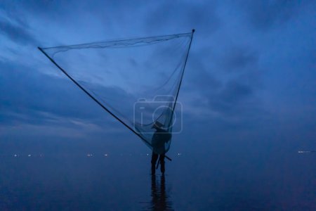Foto de Silueta de pescador vietnamita sosteniendo una red para la captura de peces de agua dulce en la naturaleza lago o río con reflexión en la mañana en Asia en Vietnam. Gente. - Imagen libre de derechos
