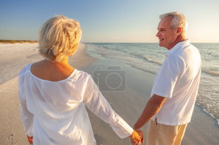 Glückliches Rentnerehepaar, das bei Sonnenuntergang am Strand spazieren geht und Händchen hält