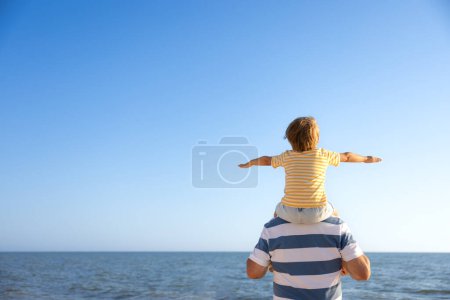 Foto de Feliz familia divirtiéndose en la playa. Padre e hijo contra el fondo azul del mar y del cielo. Vacaciones de verano y el día de los padres concepto. Retrato vista trasera - Imagen libre de derechos