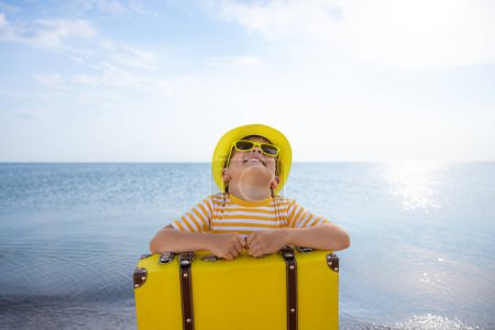 Foto de Niño feliz disfrutando del sol sobre fondo azul del cielo. Niño con maleta divirtiéndose en la playa. Vacaciones de verano y concepto de vacaciones - Imagen libre de derechos