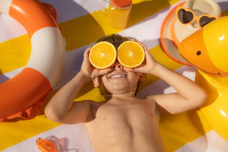 Foto de Niño feliz sosteniendo rebanadas de fruta naranja como gafas de sol. Un chico con una camiseta a rayas acostado en una toalla de playa. Comida saludable y concepto de vacaciones de verano - Imagen libre de derechos