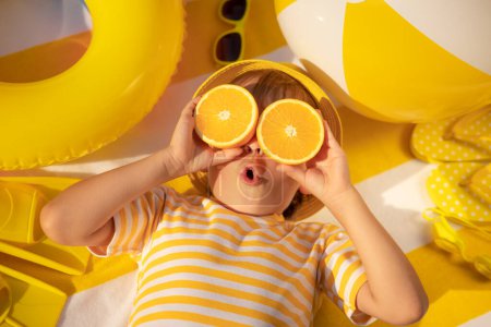 Foto de Niño sorprendido sosteniendo rebanadas de fruta naranja como gafas de sol. Un chico con una camiseta amarilla a rayas acostado en una toalla de playa. Comida saludable y concepto de vacaciones de verano - Imagen libre de derechos