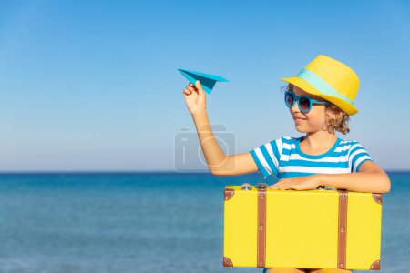 Foto de Niño con maleta vintage en vacaciones de verano. Chica divirtiéndose en la playa. Niño con avión de papel sentado sobre el fondo del mar y el cielo. Concepto de viaje y aventura - Imagen libre de derechos