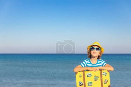 Téléchargez les photos : Enfant avec valise vintage en vacances d'été. Fille qui s'amuse sur la plage. Enfant assis sur fond de mer et de ciel. Concept de voyage et d'aventure - en image libre de droit