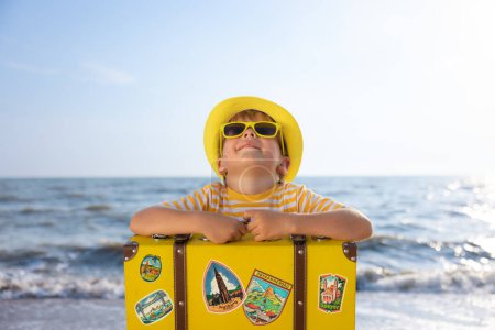 Téléchargez les photos : Joyeux enfant profitant du soleil sur fond de ciel bleu. Enfant avec valise s'amusant sur la plage. Vacances d'été et concept de vacances - en image libre de droit