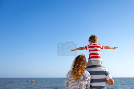 Glückliche Familie, die Spaß am Strand hat. Mutter, Vater und Kind vor blauem Meer und Himmelshintergrund. Sommerferienkonzept. Rückansicht Porträt