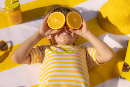 Téléchargez les photos : Heureux enfant tenant des tranches de fruits orange comme des lunettes de soleil. Un gamin portant un t-shirt rayé allongé sur une serviette de plage. Manger sainement et vacances d'été concept - en image libre de droit