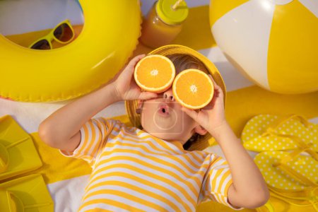Foto de Niño sorprendido sosteniendo rebanadas de fruta naranja como gafas de sol. Un chico con una camiseta amarilla a rayas acostado en una toalla de playa. Comida saludable y concepto de vacaciones de verano - Imagen libre de derechos