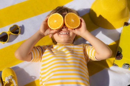 Téléchargez les photos : Heureux enfant tenant des tranches de fruits orange comme des lunettes de soleil. Un gamin portant un t-shirt rayé allongé sur une serviette de plage. Manger sainement et vacances d'été concept - en image libre de droit