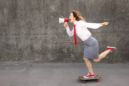 Téléchargez les photos : Heureuse femme d'affaires en skateboard. Portrait extérieur de jeune femme criant à travers haut-parleur. Retour au travail, démarrage et concept d'idée d'entreprise - en image libre de droit