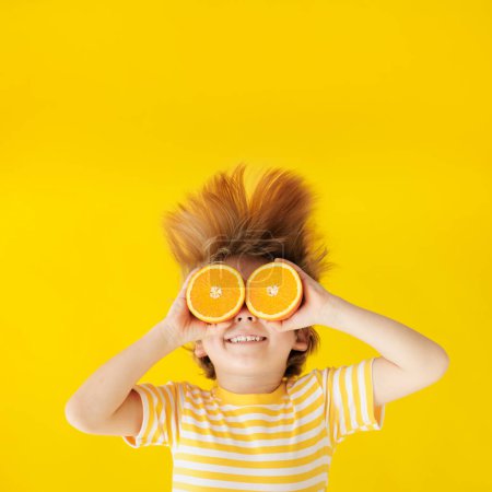 Foto de Niño sorprendido sosteniendo rebanadas de fruta naranja como gafas de sol. Niño feliz vistiendo camiseta amarilla a rayas sobre fondo de papel. Comida saludable y concepto de vacaciones de verano - Imagen libre de derechos