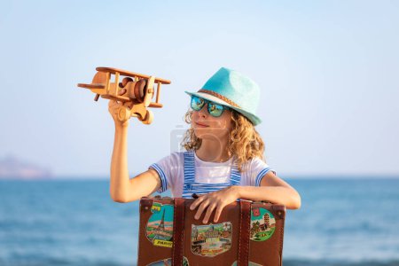 Téléchargez les photos : Enfant avec valise vintage en vacances d'été. Fille jouant avec un avion en bois sur la plage. Enfant assis sur fond de mer et de ciel. Concept de voyage et d'aventure - en image libre de droit