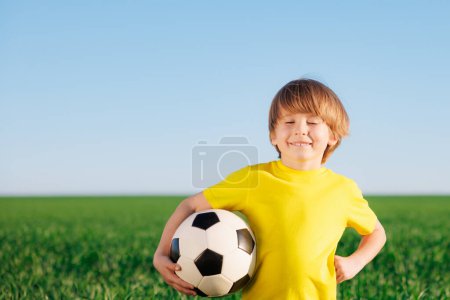 Téléchargez les photos : Un gamin heureux se fait passer pour un footballeur. Portrait d'enfant en plein air. Garçon contre champ vert. Succès et concept gagnant - en image libre de droit