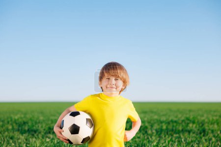 Téléchargez les photos : Un gamin heureux se fait passer pour un footballeur. Portrait d'enfant en plein air. Garçon contre champ vert. Succès et concept gagnant - en image libre de droit