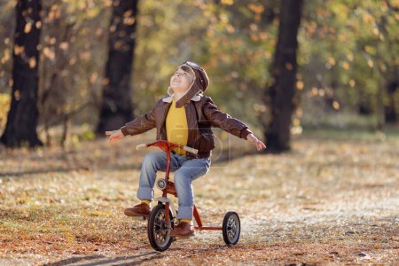 Foto de Retrato de niño feliz piloto sobre fondo de hojas amarillas. Niño divertido montar en bicicleta al aire libre en el parque de otoño. Concepto libertad e imaginación - Imagen libre de derechos