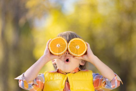 Téléchargez les photos : Étonné enfant tenant des tranches de fruits orange comme des lunettes de soleil. Enfant drôle sur fond flou automne. Joyeux garçon en automne en plein air. Concept de saine alimentation - en image libre de droit