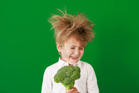 Foto de Niño sorprendido sosteniendo brócoli en clase. Chico gracioso contra el fondo de pizarra verde. Volver al concepto de escuela y educación - Imagen libre de derechos