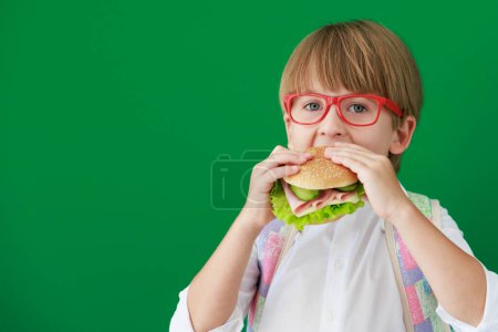 Téléchargez les photos : Enfant drôle étudiant avec hamburger en classe. Joyeux gamin contre le tableau vert. Éducation et concept de retour à l'école - en image libre de droit