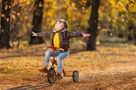 Foto de Retrato de niño feliz piloto sobre fondo de hojas amarillas. Niño divertido montar en bicicleta al aire libre en el parque de otoño. Concepto libertad e imaginación - Imagen libre de derechos