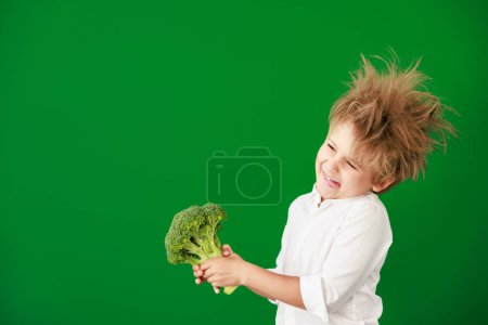 Foto de Niño sorprendido sosteniendo brócoli en clase. Chico gracioso contra el fondo de pizarra verde. Volver al concepto de escuela y educación - Imagen libre de derechos