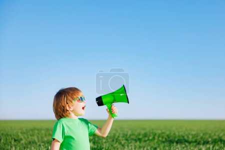 Téléchargez les photos : Joyeux enfant criant à travers haut-parleur sur fond bleu ciel d'été. Enfant s'amuser dans le champ vert de printemps en plein air. Concept du Jour de la Terre - en image libre de droit