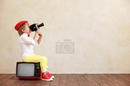 Téléchargez les photos : Garçon drôle tenant caméra vintage sur fond de mur grunge. Un gamin assis sur une vieille télé. Enfant jouant à la maison - en image libre de droit