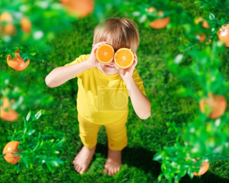 Foto de Niño sorprendido sosteniendo rebanadas de fruta naranja como gafas de sol. Niño vistiendo camiseta amarilla de pie sobre hierba verde. Comida saludable y concepto de vacaciones de verano - Imagen libre de derechos