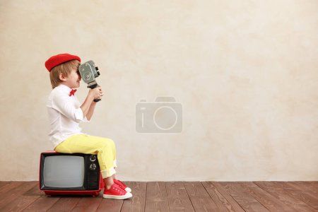 Foto de Chico divertido sosteniendo cámara vintage contra fondo de pared grunge. Un chico sentado en la vieja tele. Niño jugando en casa - Imagen libre de derechos