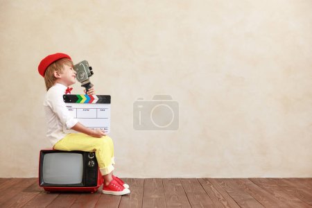 Téléchargez les photos : Garçon drôle tenant caméra vintage et claquette sur fond de mur grunge. Un gamin assis sur une vieille télé. Enfant jouant à la maison - en image libre de droit