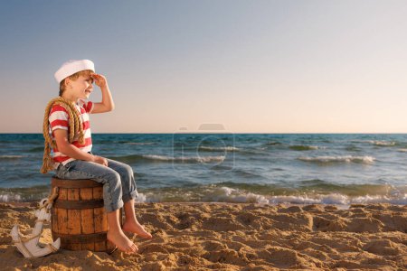 Foto de El niño finge ser marinero. Un chico sentado en un barril viejo en la playa. Un chico de vacaciones de verano. Concepto de aventura y viajes - Imagen libre de derechos