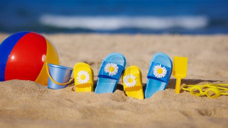 Foto de Chanclas para niños, pelota de playa y snorkel sobre arena amarilla sobre fondo azul marino. Concepto vacaciones de verano - Imagen libre de derechos
