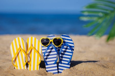 Photo for Flip-flops and sunglasses on yellow sand against blue sea background. Summer vacation and travel concept - Royalty Free Image