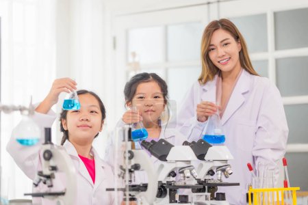 Foto de Clase de ciencias primarias, Niños alegres con el profesor científico mostrando botella de prueba con líquido químico en el laboratorio de la escuela, Laboratorio de ciencias - Imagen libre de derechos
