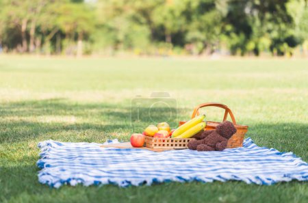 Foto de Cesta de picnic con frutas y muñecas en tela azul en el jardín - Imagen libre de derechos