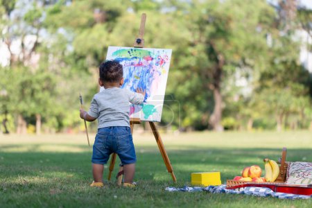 Téléchargez les photos : Vue arrière de la peinture de garçon enfant sur toile dans le jardin, enfant heureux garçon dessin une image en plein air - en image libre de droit