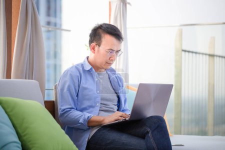 Téléchargez les photos : Asiatique homme utilisant ordinateur portable assis sur canapé à la maison, Homme travaillant à partir d'un ordinateur portable à la terrasse - en image libre de droit
