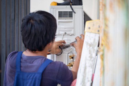Foto de Joven reparador que fija la unidad del acondicionador de aire, hombre técnico asiático que instala un aire acondicionado en una casa del cliente, conceptos de mantenimiento y reparación - Imagen libre de derechos