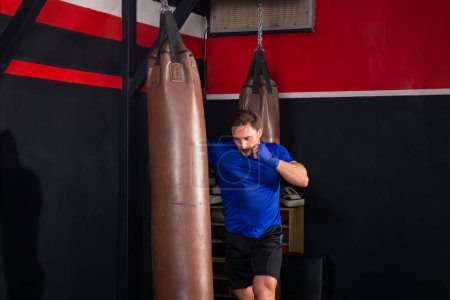 Téléchargez les photos : Boxeur homme exercices coups de poing avec sac de boxe dans la salle de gym, Boxer frapper un énorme sac de boxe dans un studio de boxe - en image libre de droit