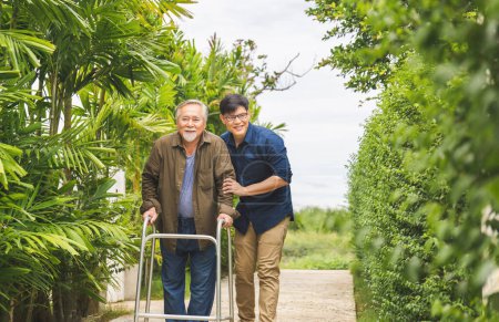 Foto de Cuidador está enseñando anciano a caminar con andador, padre asiático mayor y de mediana edad hijo caminar en el parque, anciano hombre mayor con hijo caminar al aire libre - Imagen libre de derechos
