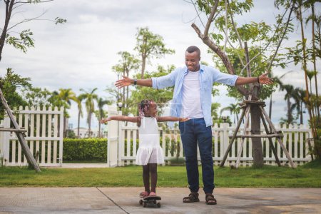Téléchargez les photos : Père aidant sa petite fille à monter sur un skateboard, Petite fille et son père jouant avec le skateboard à l'extérieur, concepts de la fête des pères - en image libre de droit