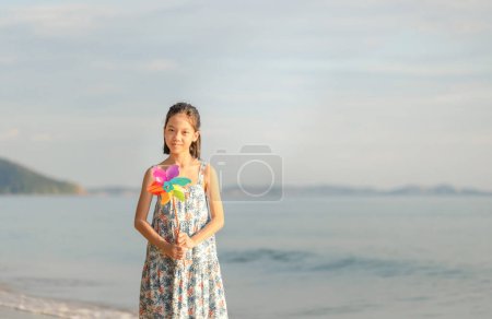 Photo for Portrait of Happy little Asian child girl playing on the beach holds windmill in hand, Kid playing outdoors with pinwheels - Royalty Free Image