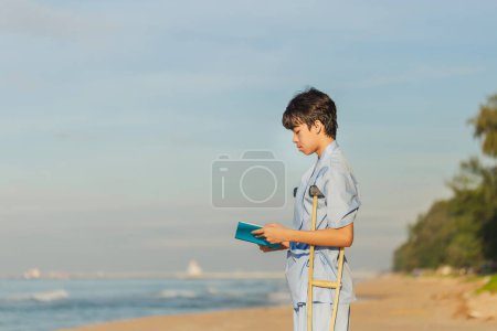 Téléchargez les photos : Jeunes patients avec des béquilles lisant un livre sur la plage le matin, Homme avec des béquilles en voyage sur la plage. - en image libre de droit