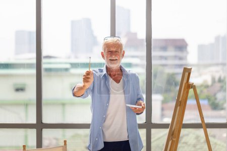 Foto de Hombre mayor alegre mostrando pincel y pintura sobre lienzo en casa, Hombre mayor pintando sobre lienzo, Conceptos de jubilación feliz - Imagen libre de derechos