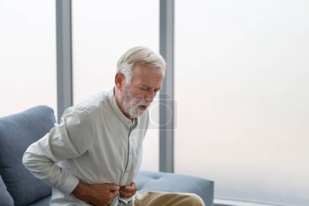 Foto de Hombre mayor infeliz con dolor de estómago, el hombre presiona su mano al estómago de dolor insoportable, hombre maduro con dolor de estómago sensación de malestar sentado en la sala de estar - Imagen libre de derechos