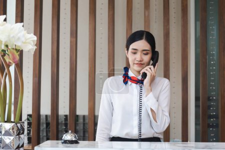 Téléchargez les photos : Jeune femme asiatique réceptionniste derrière le comptoir de l'hôtel parlant au téléphone, Femme réceptionniste travaillant dans l'hôtel - en image libre de droit