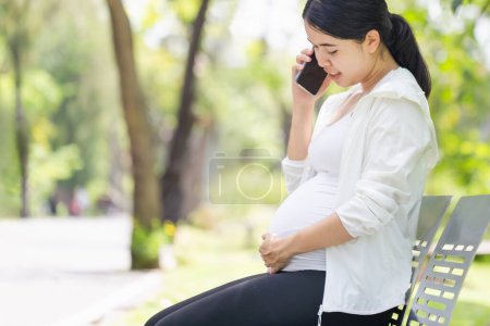 Foto de Mujer embarazada sentada relajada en el parque y usando un smartphone - Imagen libre de derechos
