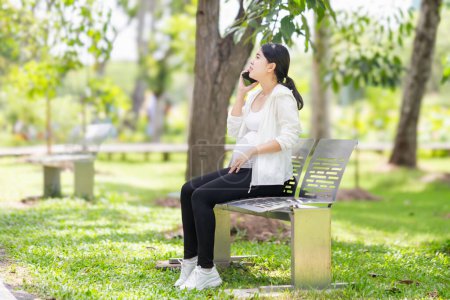 Foto de Mujer embarazada sentada relajada en el parque y usando un smartphone - Imagen libre de derechos