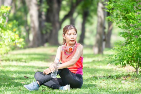 Téléchargez les photos : Athlète jeune belle femme avec une bouteille d'eau en plastique et écouteurs au parc vert, Sportswoman boire de l'eau après l'exercice - en image libre de droit